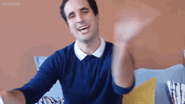 a man in a blue sweater is sitting on a couch and waving his hand .