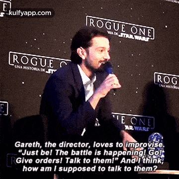 a man is talking into a microphone while sitting in front of a sign that says `` rogue one '' .