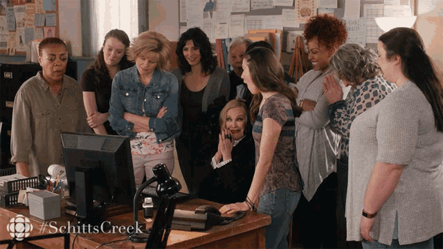 a group of women are standing around a desk with a computer monitor and the words schitts creek on the bottom right