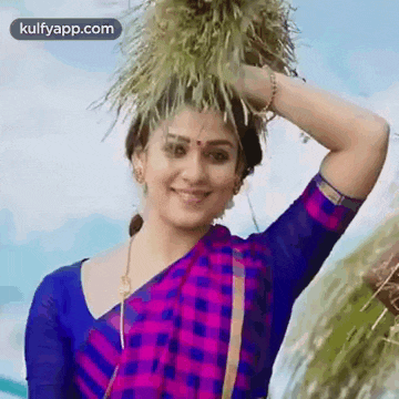 a woman in a purple and blue saree is holding a bunch of grass on her head .