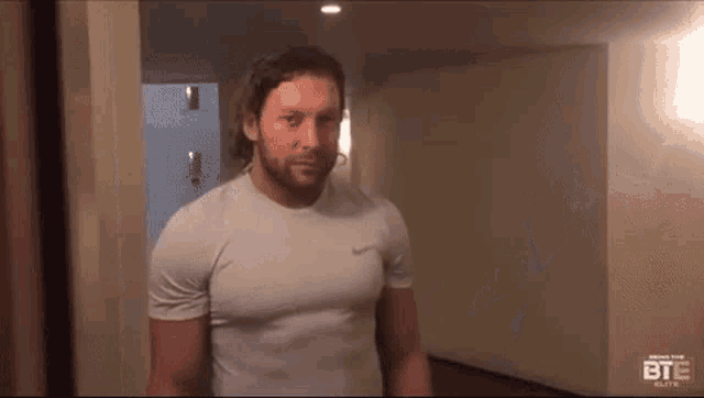 a man with a beard is wearing a white nike shirt and standing in a hallway .