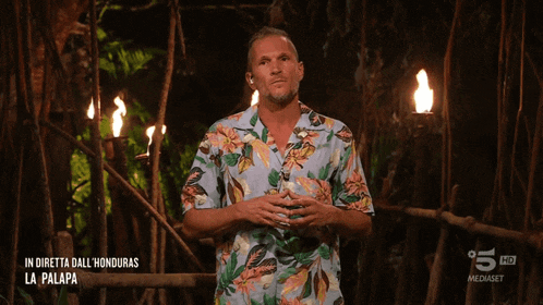 a man in a hawaiian shirt stands in front of a fire in a jungle