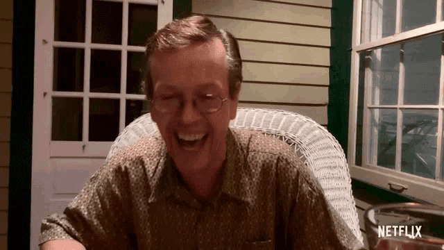 a man sitting in a wicker chair laughing with a netflix logo in the background