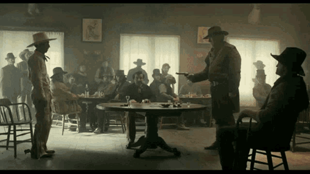 a group of men in cowboy hats are gathered around a table with a man pointing a gun