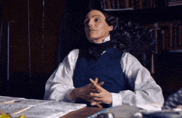 a man in a blue vest and tie is sitting at a desk with his hands folded .
