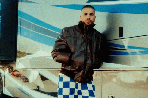 a man wearing a leather jacket and plaid pants leans against a blue and white bus