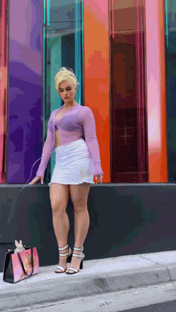 a woman wearing a purple top and a white skirt is standing on the sidewalk .