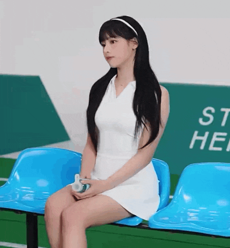 a woman in a white dress is sitting on a blue bench holding a bottle .