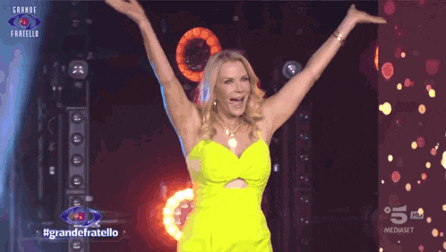a woman in a neon yellow dress stands on a stage with her arms in the air in front of a sign that says grande fratello