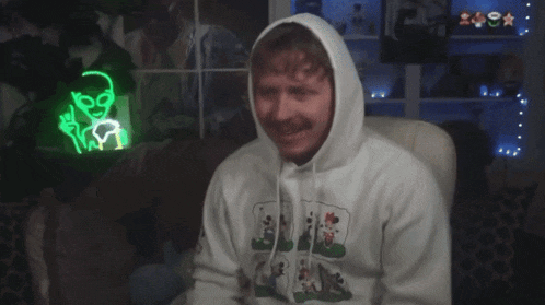 a man wearing a white hoodie is sitting in front of a neon sign that says " alien "
