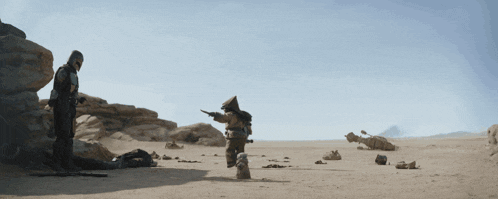 a man standing in the desert holding a gun with another man standing behind him