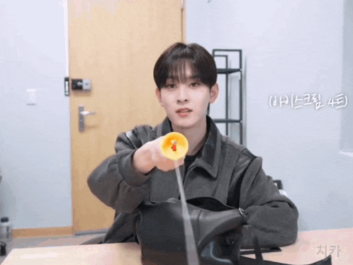 a young man is sitting at a table holding a yellow object and a black bag with korean writing on it