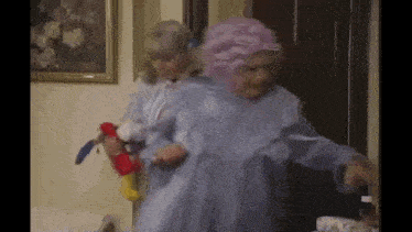 two women are standing next to each other in a room . one of the women is wearing a pink wig .