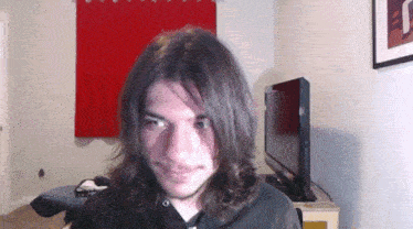 a man with long hair looks at the camera in front of a tv