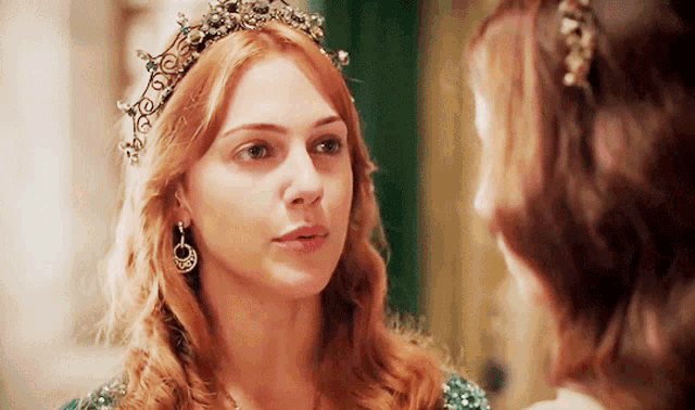 a woman with red hair wearing a tiara looks at another woman
