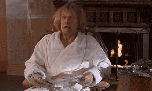a man in a bathrobe is sitting in front of a fireplace smoking a cigarette and reading a newspaper .