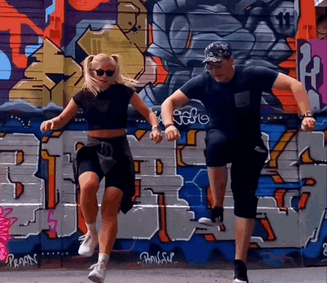 a man and a woman are jumping in front of a graffiti wall that has the letters t and t on it
