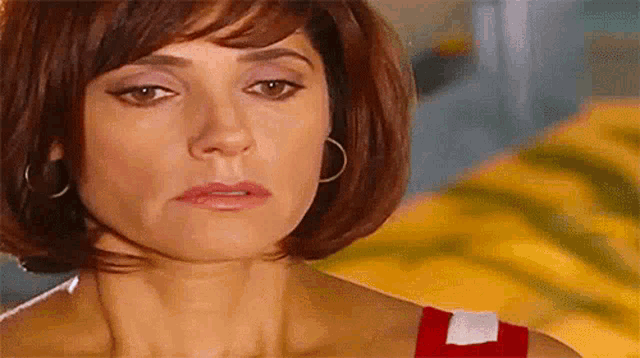 a woman with short red hair and hoop earrings is looking at the camera with a sad look on her face .