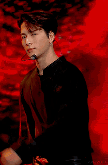 a man with a microphone on his ear stands in front of a red background that says axtzzles
