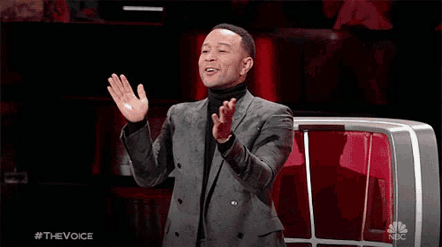 a man in a suit and turtleneck applauds in front of a nbc television