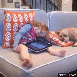 a little girl is laying on a couch looking at a tablet while a dog looks on .
