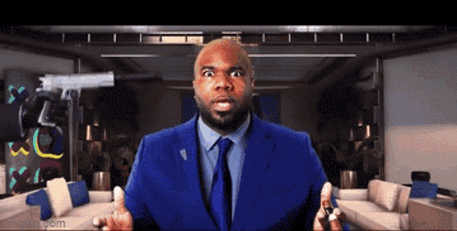 a man in a blue suit and tie is standing in a living room with a gun behind him