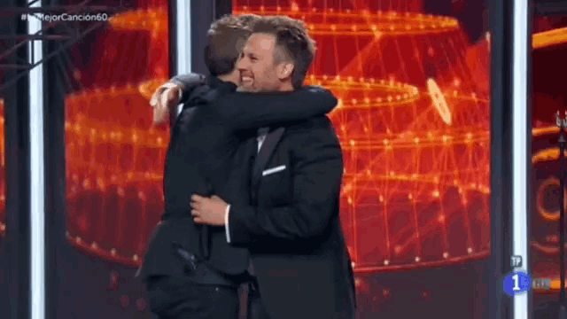 a man in a suit is hugging another man in a tuxedo on a stage