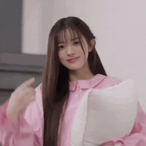 a woman in a pink shirt is holding a white pillow and giving a thumbs up .