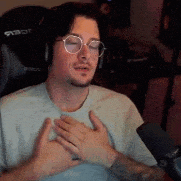 a man wearing glasses and headphones is sitting in front of a microphone and holding his chest .