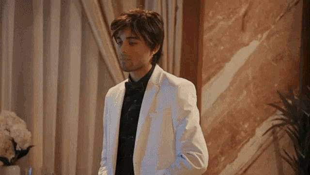 a man in a white tuxedo and bow tie is standing in front of a marble wall .