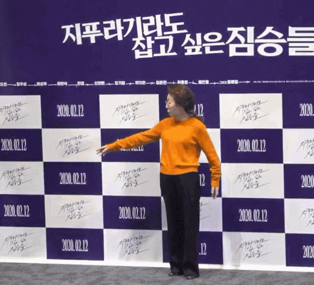 a woman in an orange sweater stands in front of a backdrop that says 2020 on it
