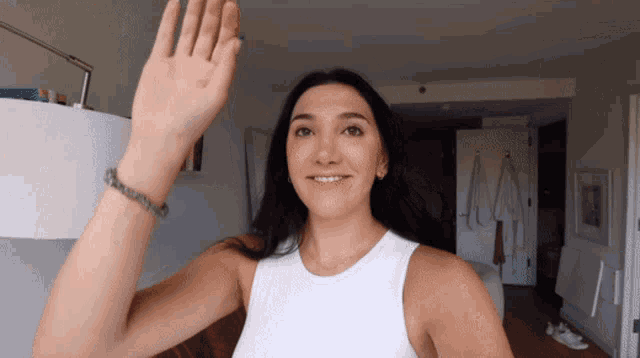 a woman in a white tank top is smiling and waving her hand