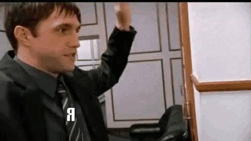 a man in a suit and tie is standing in front of a door and waving his hand .
