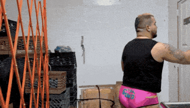 a man in a black tank top and pink underwear with the word bomb on the back is standing in front of a stack of crates