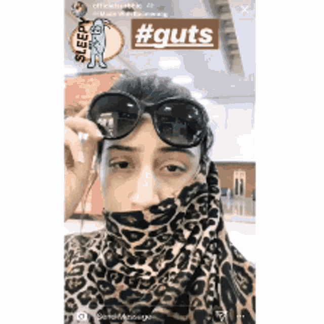 a woman wearing sunglasses and a leopard print scarf has a sticker on her face that says #guts