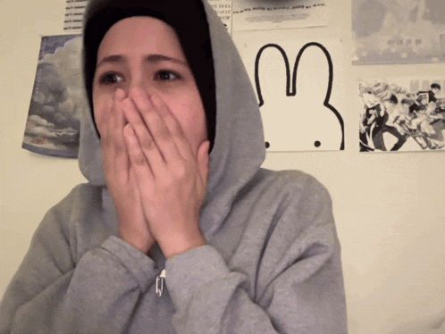 a person wearing a hoodie covering their mouth in front of a wall with a drawing of a rabbit on it
