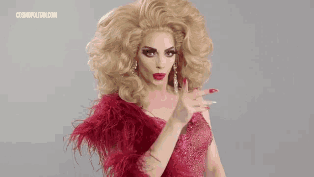 a drag queen is wearing a red dress and feathers and is holding her finger to her lips .