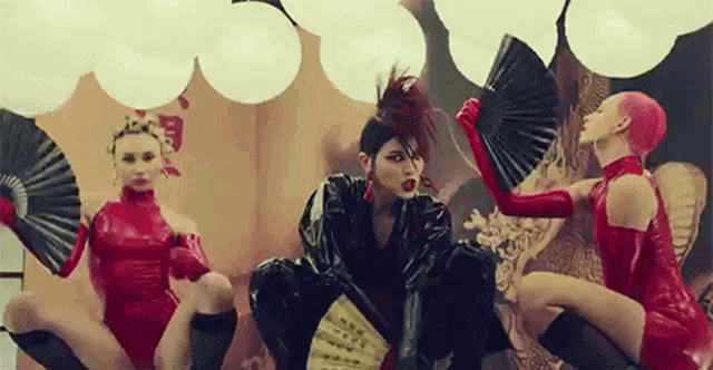 a woman in a black leather jacket is holding a fan while two other women in red latex dresses are holding fans .