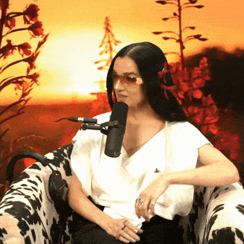 a woman wearing sunglasses sits in a chair with a microphone in front of her