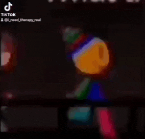 a clown is standing in the dark with a microphone in his hand and a rainbow colored hat on .