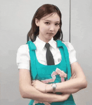 a woman in a blue apron and tie is standing with her arms crossed and looking at the camera .