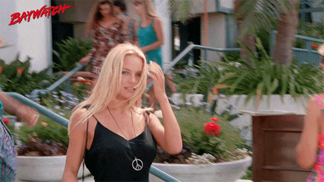 a woman wearing a black tank top and a peace sign necklace stands in front of a baywatch sign