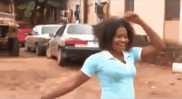 a woman in a blue shirt is dancing in a parking lot with cars in the background .
