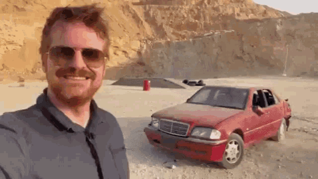 a man wearing sunglasses is taking a selfie next to a red car in the desert .