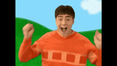 a man in an orange sweater is standing in front of a green hill with his arms in the air .