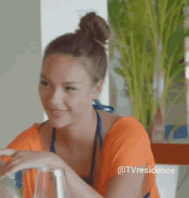 a woman in an orange shirt is sitting at a table with a plant in the background and the caption tvresidence