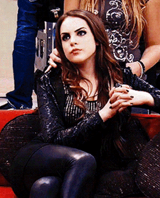 a woman in a black jacket sits on a couch with her hands folded