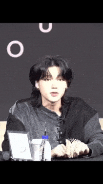 a young man with long black hair is sitting at a table with a bottle of water