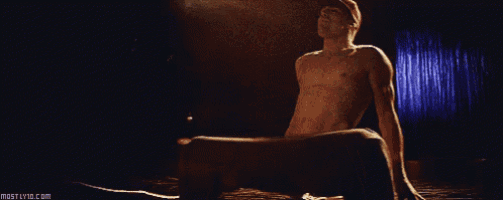 a man without a shirt is sitting on a stage with the website mostlyvid.com in the lower right corner