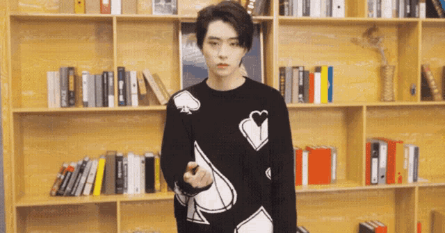 a man wearing a black sweater with spades and hearts on it stands in front of a bookshelf